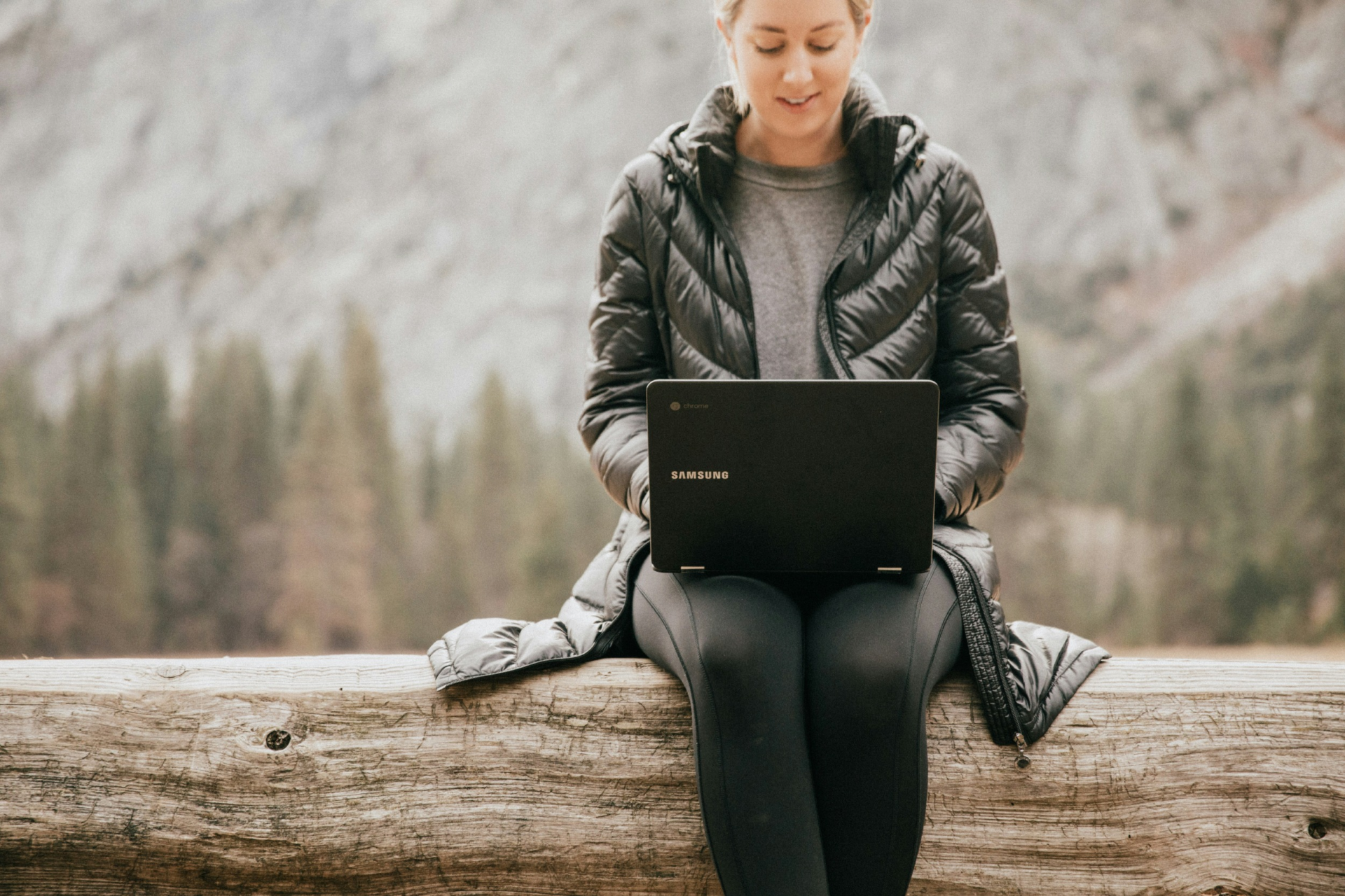 A lady in puffer jacket sits on a log outdoors, reading from her laptop